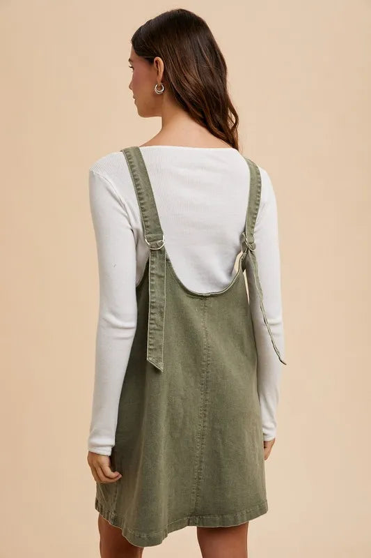 Army green Annie Wear denim overall dress with V-neck, adjustable straps, and front pockets, styled with rolled cuffs.
