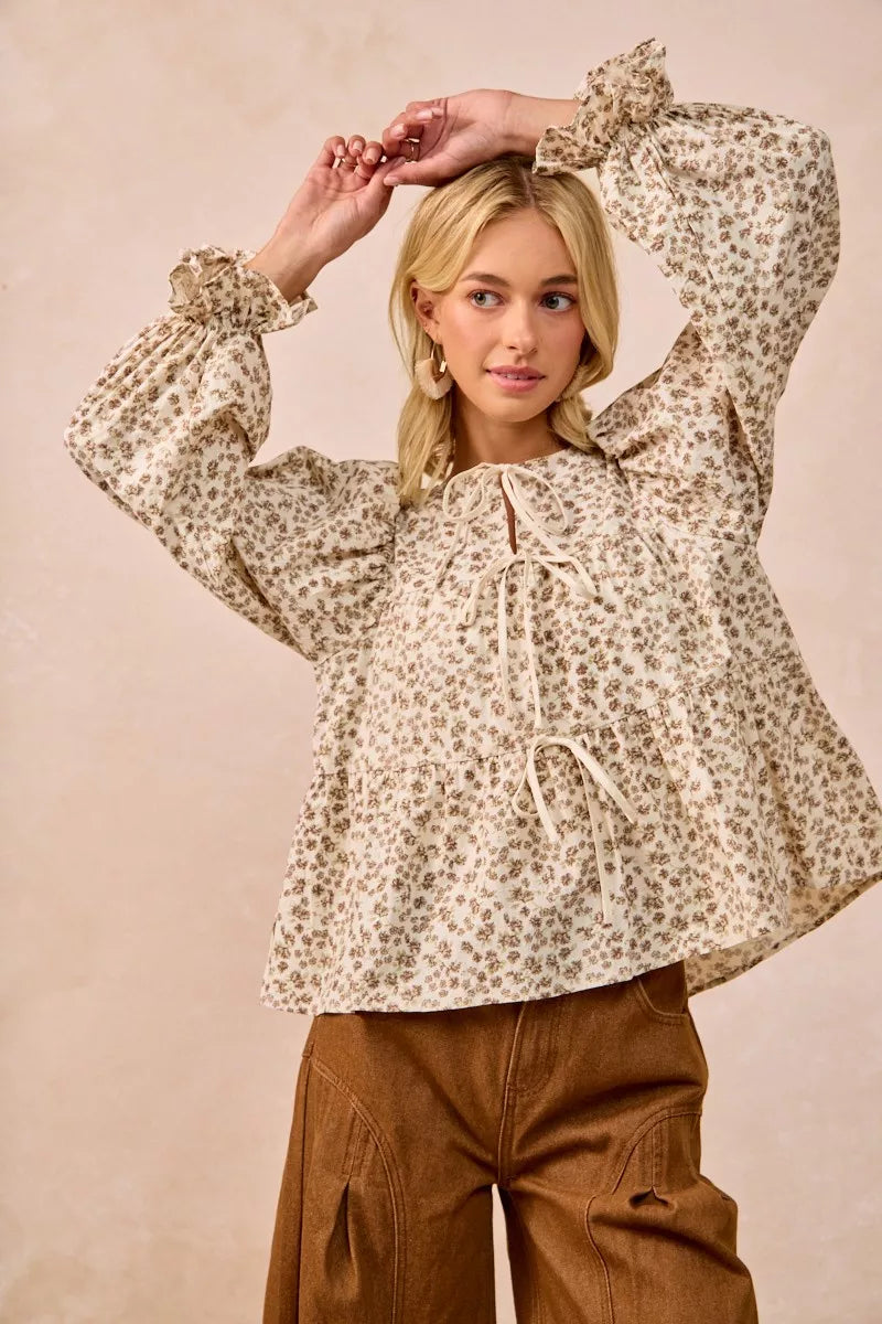 Oatmeal BiBi floral printed peplum blouse by Trendsi with shirring and tied detail, displayed on a hanger.