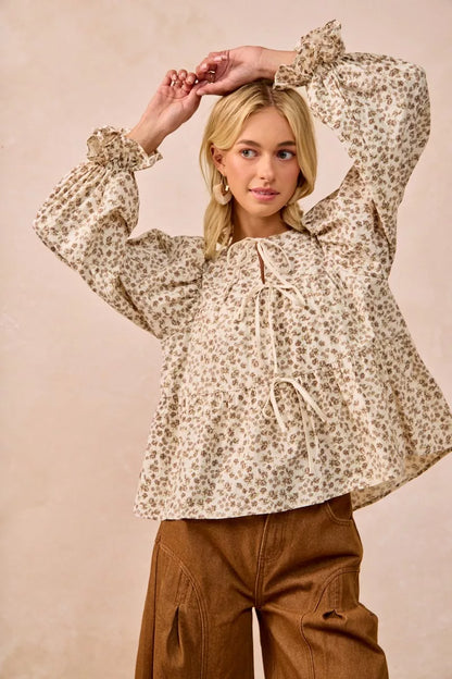 Oatmeal BiBi floral printed peplum blouse by Trendsi with shirring and tied detail, displayed on a hanger.